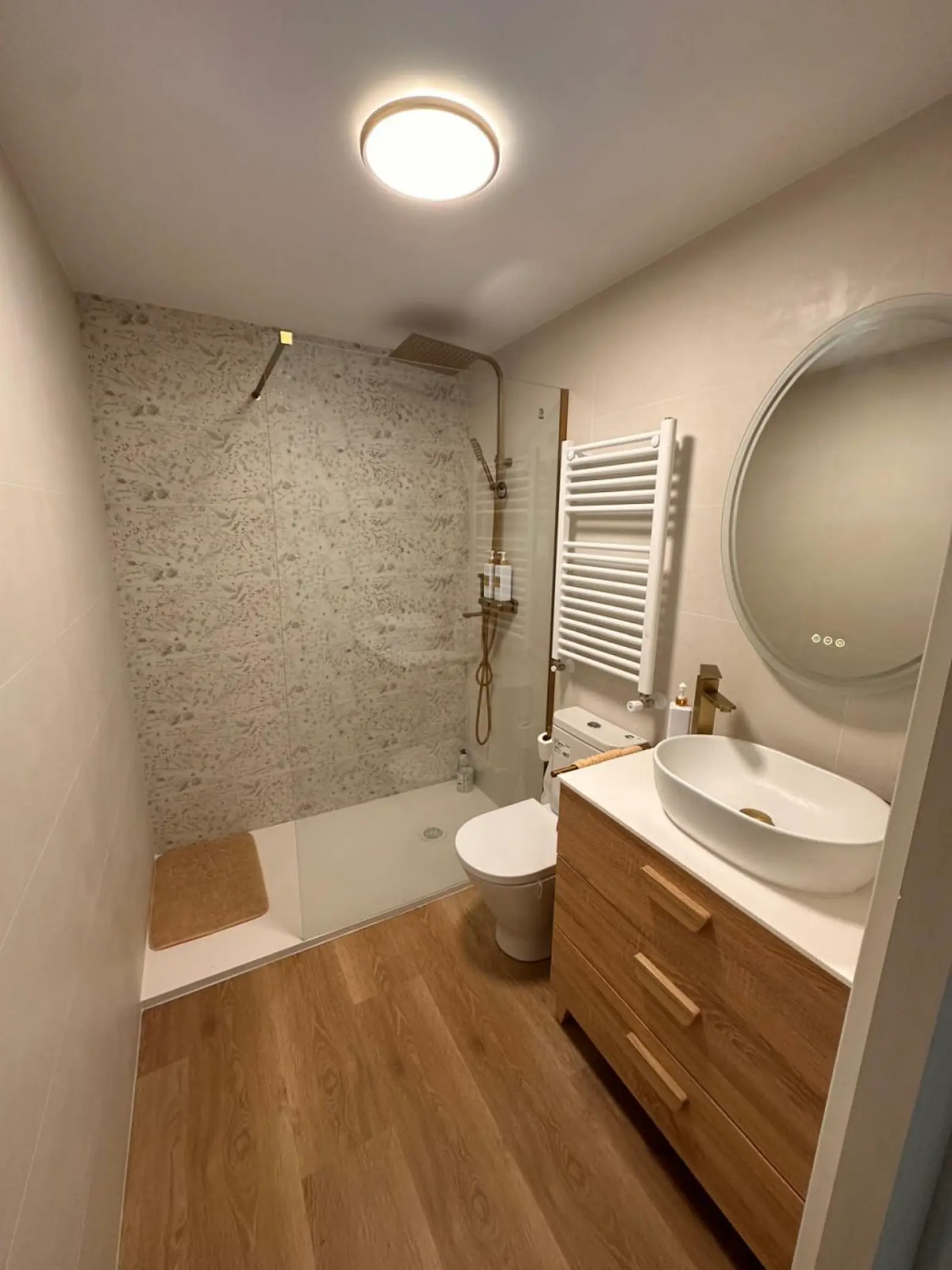 Baño reformado con plato de ducha, revestimiento claro, radiador toallero y mueble de madera